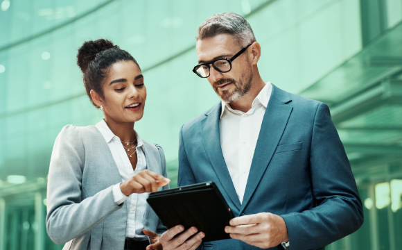Two commercial real estate pros, one female one male, look over an iPad while standing in front of a heavily windowed building. 