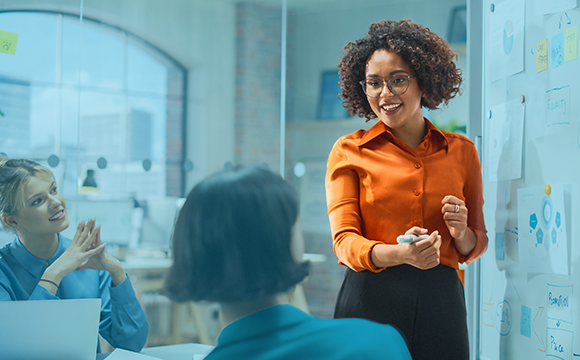 Female lease administration professional with curly hair, glasses and orange shirt presents at a whiteboard.